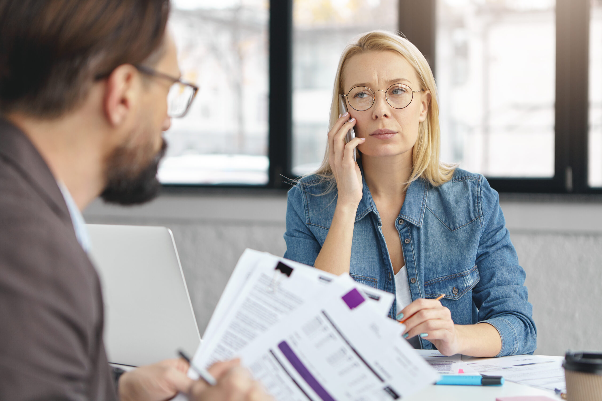 Attractive middle aged blonde female manger in spectacles calls to colleagues, organizes business meeting to discuss main issues of work, looks at HOALiving Community Mnaager who studies documents sits back to camera